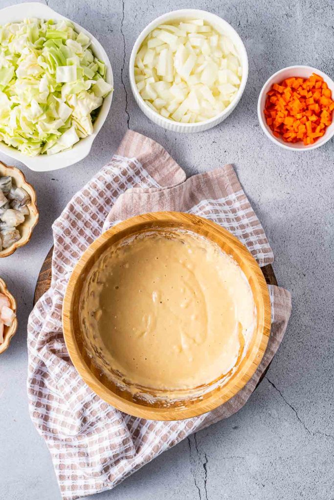 A wooden bowl of batter sits on a kitchen towel, surrounded by bowls of chopped cabbage, onions, carrots, and shrimp on a gray countertop.