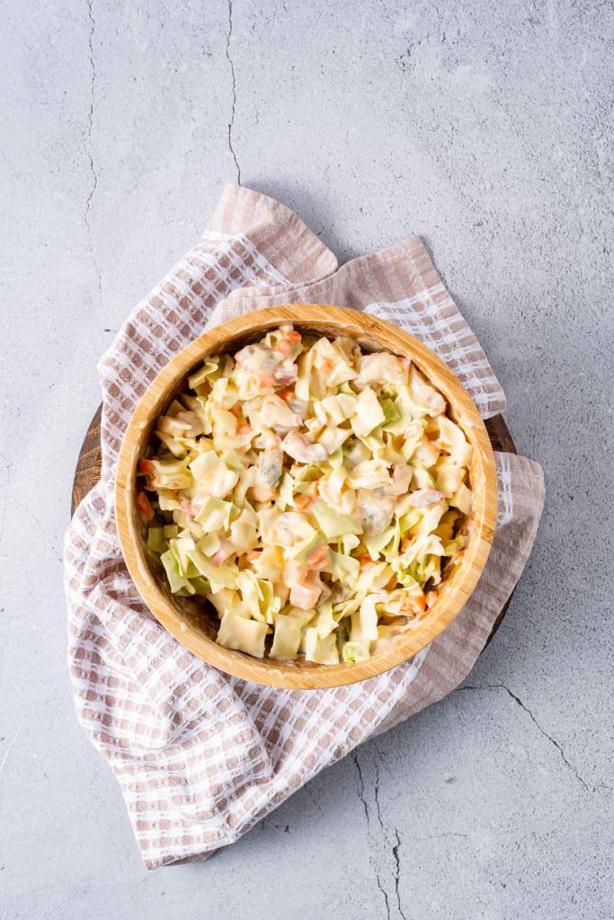 A wooden bowl filled with creamy coleslaw sits on a checkered cloth atop a light gray, textured surface.