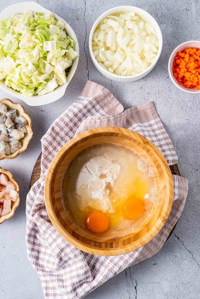 A wooden bowl with eggs, flour, and water sits on a towel, surrounded by bowls of chopped cabbage, onion, carrots, shrimp, and bacon on a gray surface.