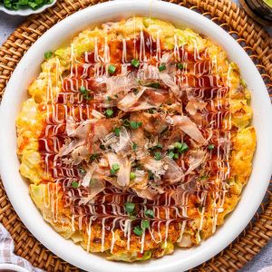 A plate of okonomiyaki topped with mayonnaise, okonomiyaki sauce, bonito flakes, and chopped green onions, served on a white dish.