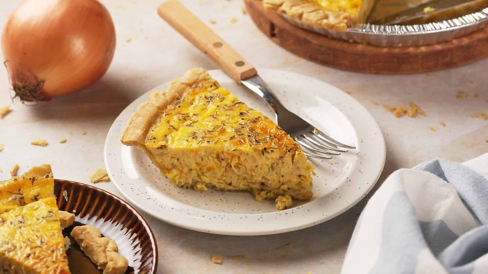 A slice of quiche on a white plate with a fork, with a whole onion, a pie dish, and a napkin nearby.