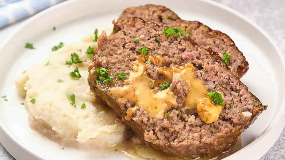 Two slices of meatloaf with melted cheese inside, served next to mashed potatoes and garnished with chopped parsley on a white plate.