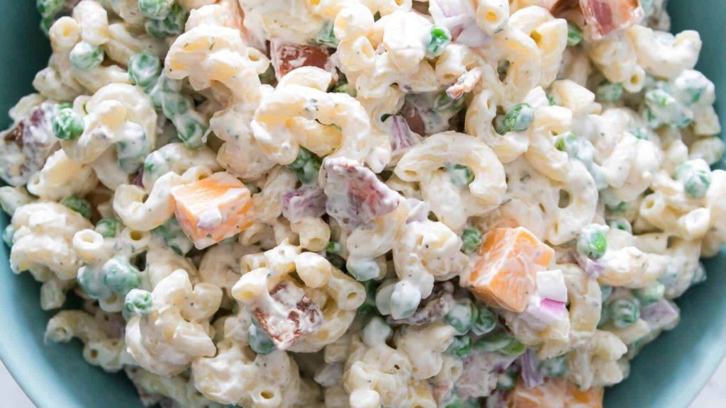 Close-up of macaroni salad with elbow pasta, peas, cheddar cheese cubes, red onion, bacon pieces, and creamy dressing.
