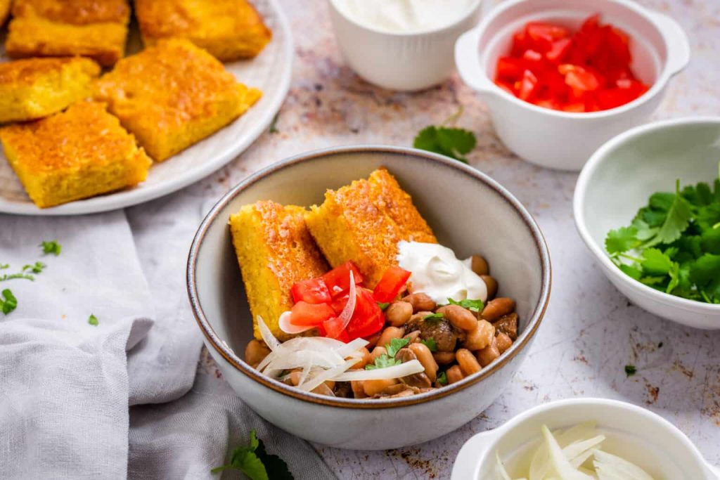 A bowl with pinto beans, sliced onions, chopped tomatoes, sour cream, and two pieces of cornbread—classic recipes come alive here. Surrounding dishes offer extra cornbread, tomatoes, onions, and cilantro for an authentic touch.