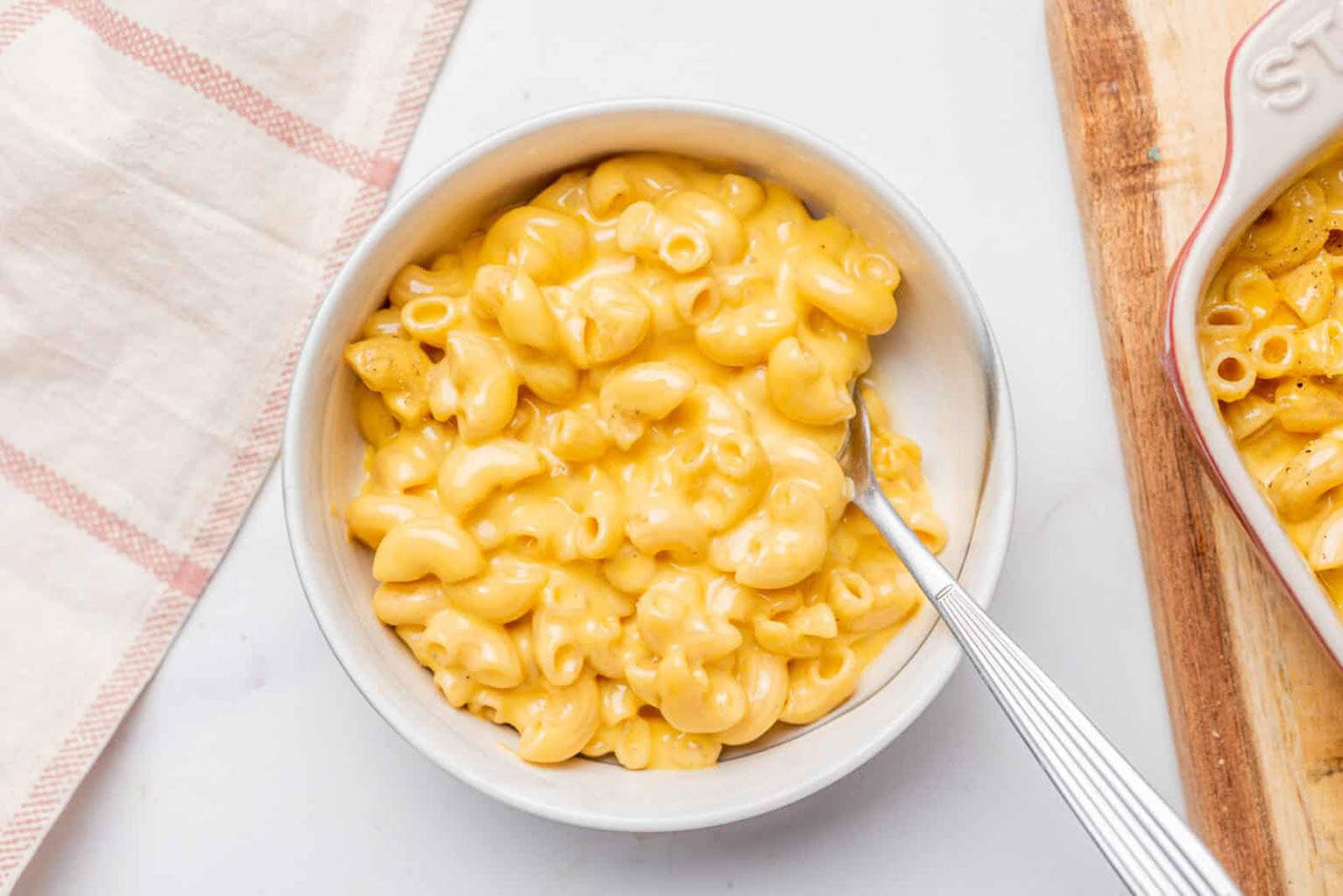 A bowl of creamy macaroni and cheese with a fork, placed on a white surface next to a beige-striped cloth and a wooden board.