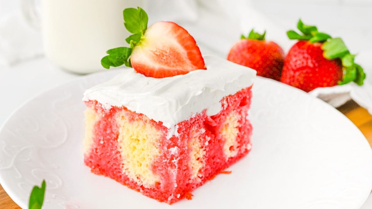 A slice of strawberry poke cake with white whipped cream topping and a halved strawberry on top, served on a white plate. Whole strawberries and a glass of milk are in the background.