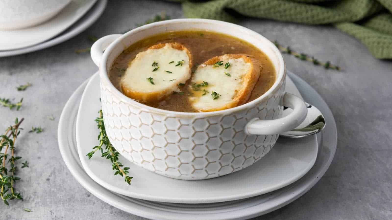 A bowl of French onion soup topped with two slices of toasted bread and melted cheese, garnished with fresh thyme, served on a white plate.