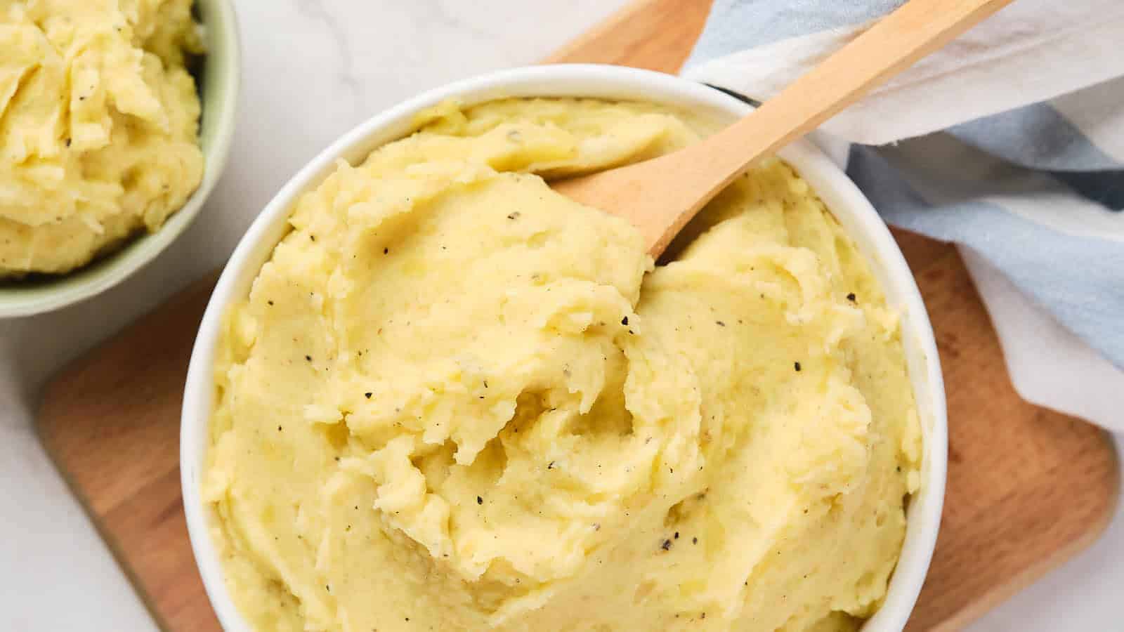 A bowl of creamy mashed potatoes with black pepper, served with a wooden spoon on a cutting board.