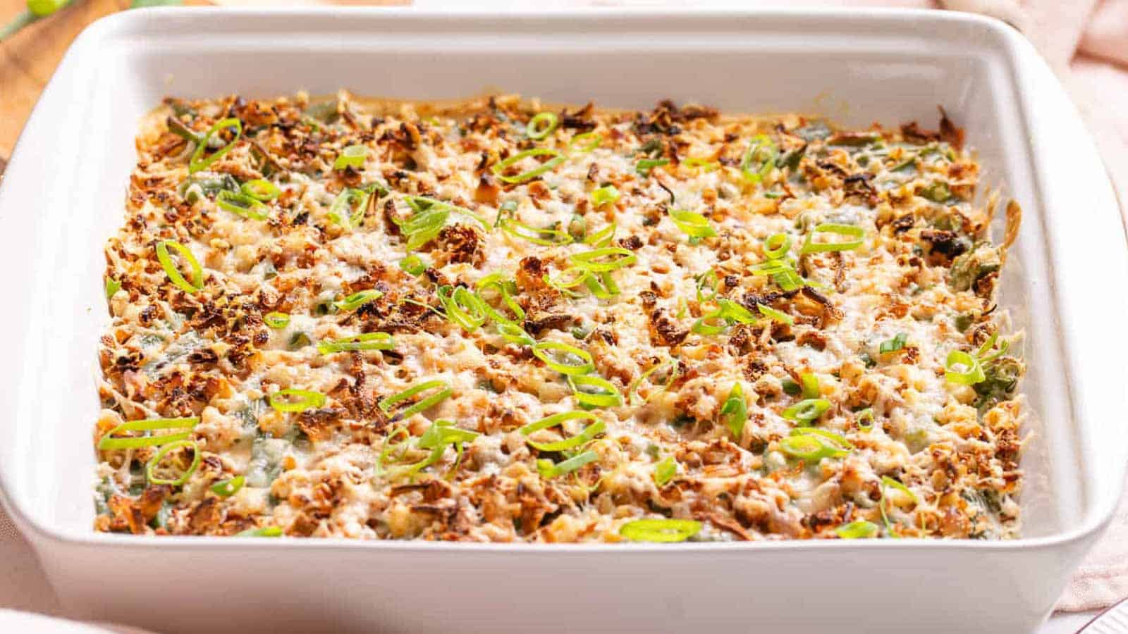 A rectangular white baking dish filled with a baked casserole topped with melted cheese, browned bits, and chopped green onions.