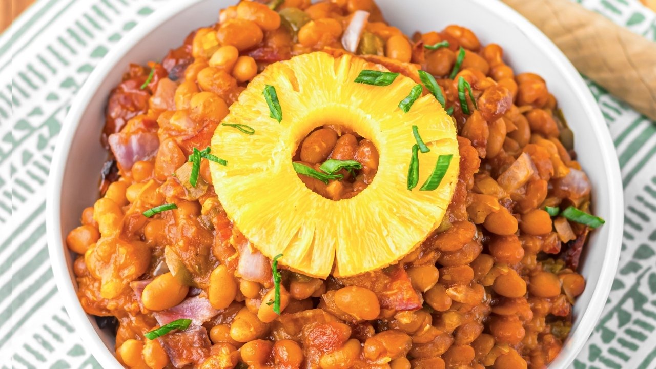 A bowl of Hawaiian Baked Beans garnished with pineapple sits on a striped cloth.