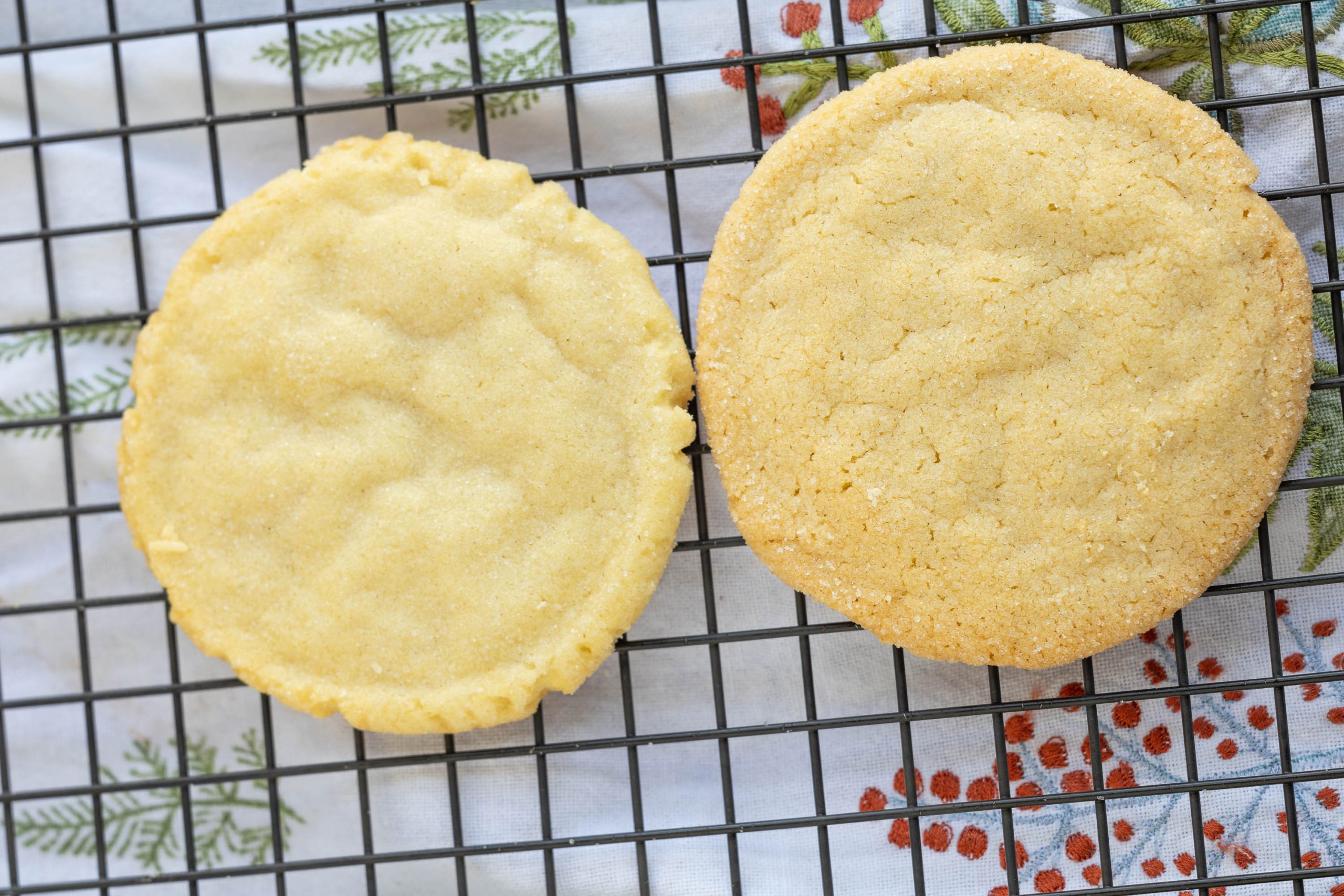 Two sugar cookies cooling on a black wire rack over a white cloth with green fern and red berry patterns.