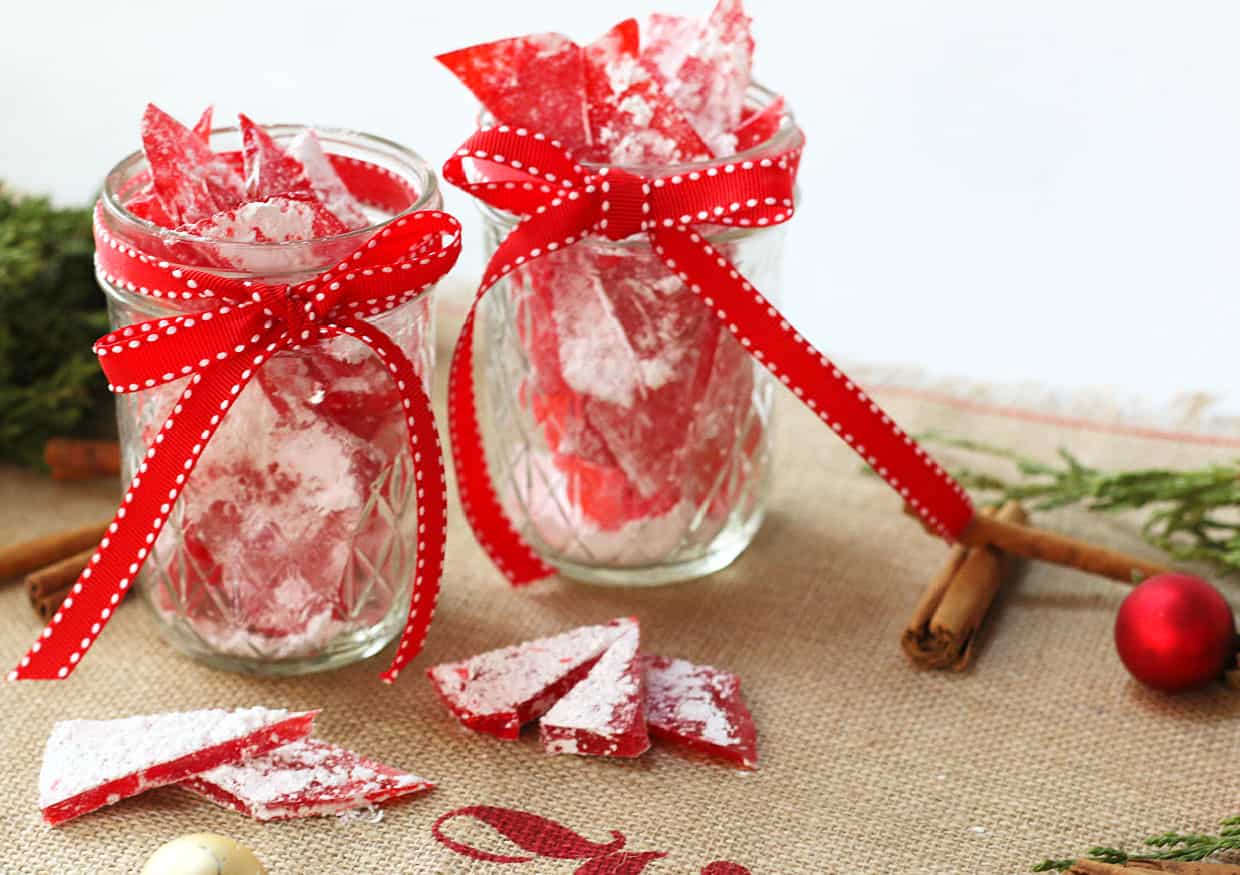 Two glass jars filled with red peppermint candy bark, tied with red ribbons, are displayed on a burlap surface with cinnamon sticks, greenery, and holiday decorations.