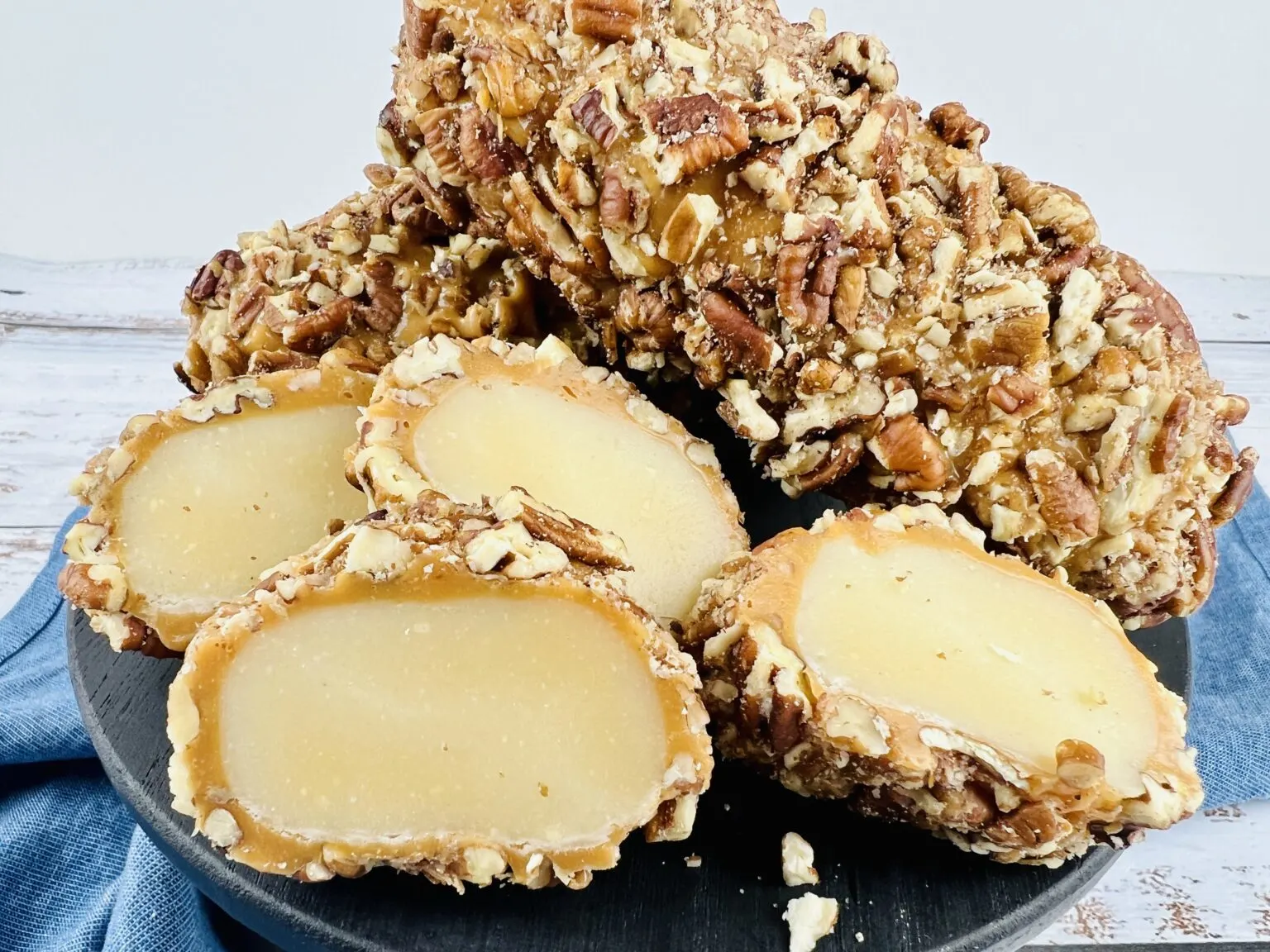 A black plate with whole and sliced pecan-coated logs filled with a dense, light-colored center, displayed on a light surface with a blue cloth underneath.
