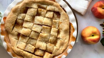 A baked peach pie with a lattice crust in a white pie dish, with two fresh peaches and a folded napkin on a marble surface nearby.