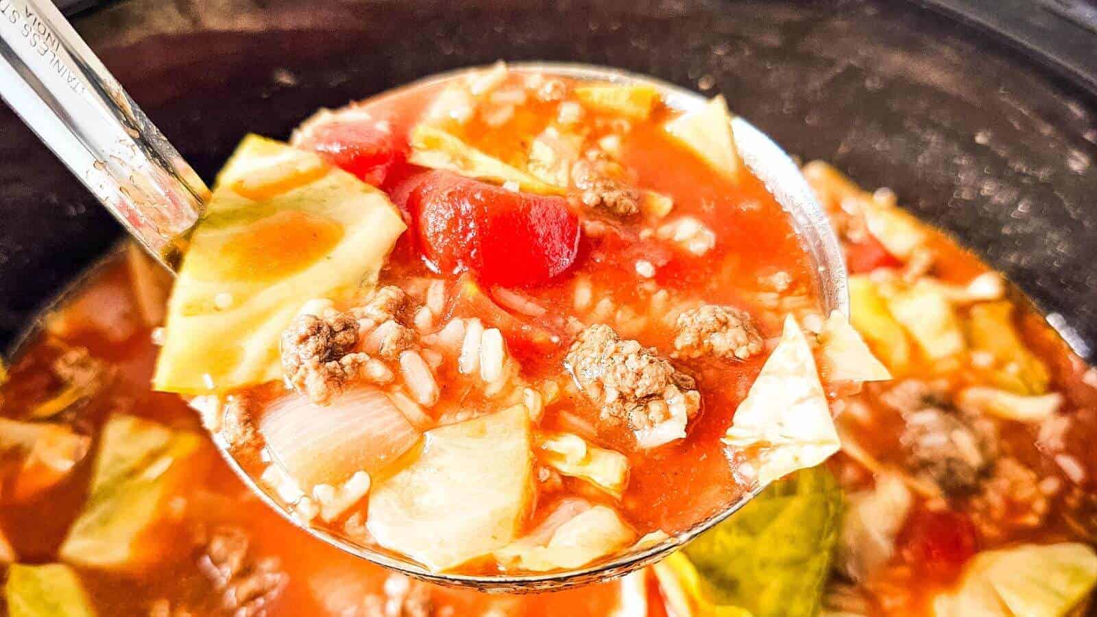 Close-up of a ladle holding a hearty soup with visible chunks of tomato, cabbage, ground meat, and grains in broth.