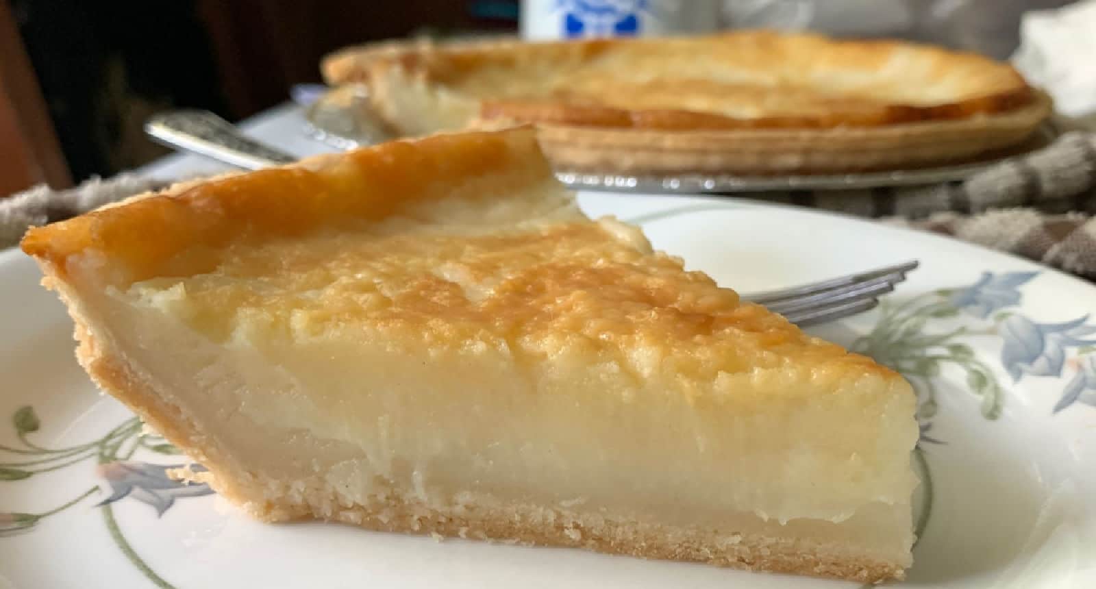 A close-up of a slice of custard pie on a decorative plate, with the rest of the pie visible in the background.