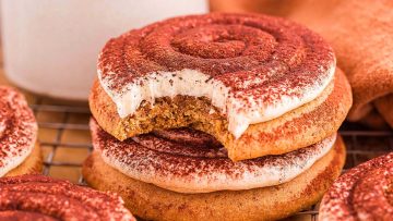 A stack of frosted cookies dusted with cocoa powder sits on a cooling rack, with the top cookie showing a visible bite.