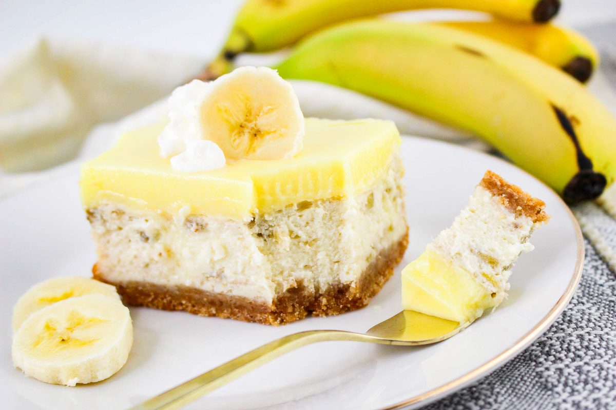 A slice of banana pudding cheesecake with a graham cracker crust sits on a plate, garnished with a banana slice and whipped cream; bananas and cheesecake squares are in the background.