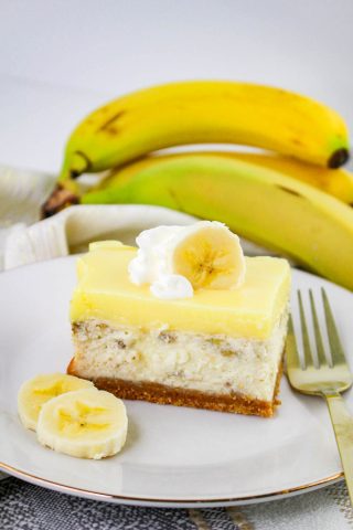 A slice of Banana Pudding Cheesecake topped with whipped cream and a banana slice, served on a white plate with banana slices, a fork, and ripe bananas in the background.