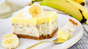A slice of banana cream cheesecake with a graham cracker crust, topped with banana slices and whipped cream, is served on a plate with a fork—perfect for fans of classic cheesecake or banana pudding treats. Whole bananas sit in the background.