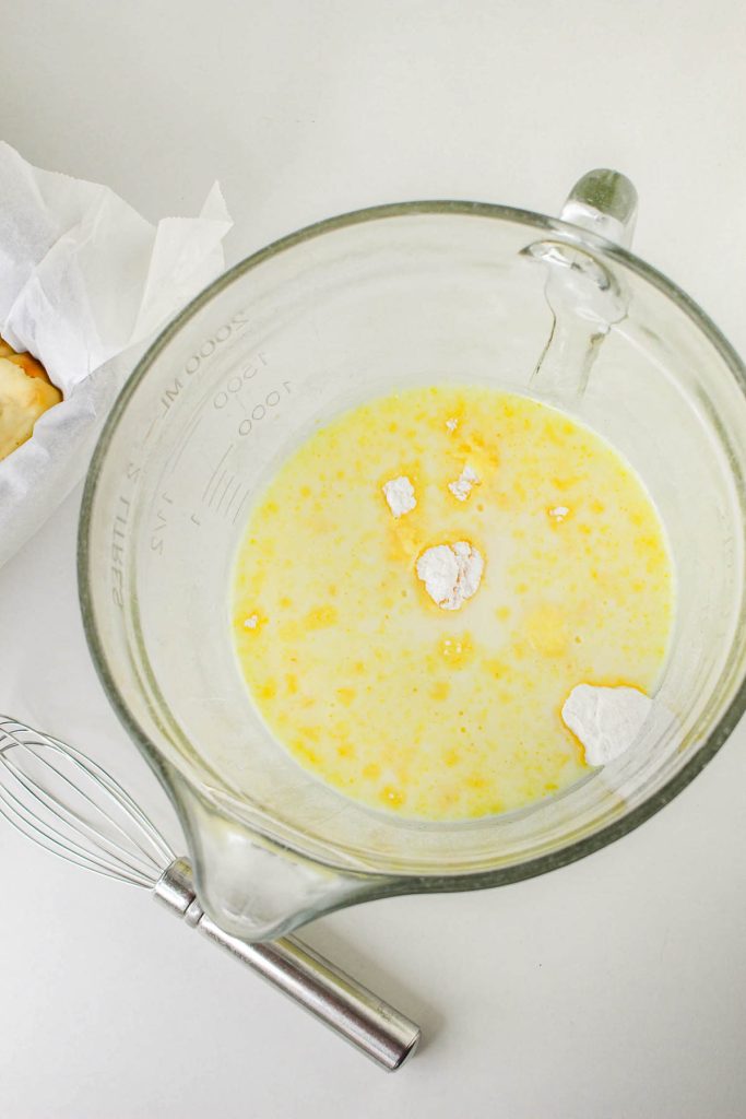 A glass measuring cup containing milk, eggs, and flour being mixed, with a whisk and parchment paper nearby—ingredients coming together for delicious Banana Pudding Cheesecake on a white surface.
