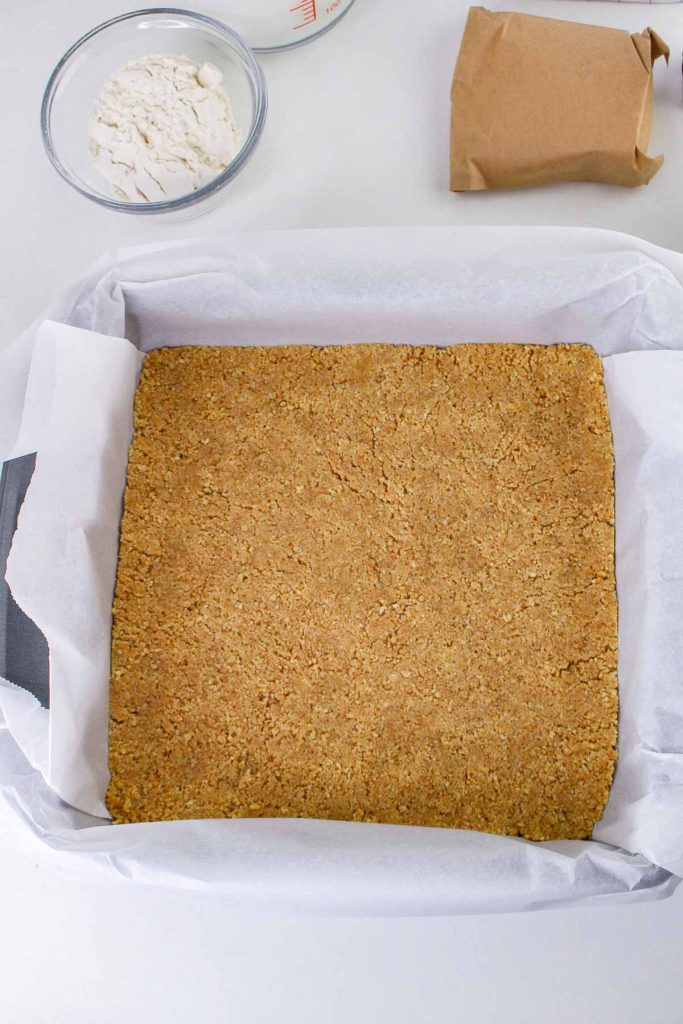 A square baking pan lined with parchment paper holds a pressed graham cracker crust, ready to become delicious Banana Pudding Cheesecake Squares; a bowl of flour and a brown paper bag sit nearby on the countertop.