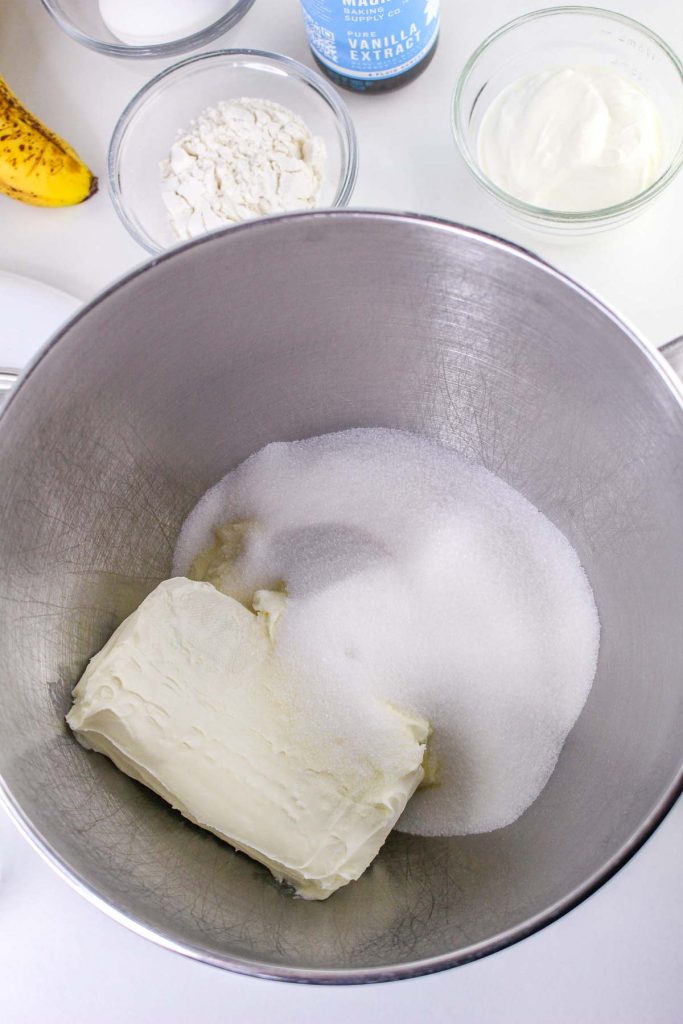 A metal mixing bowl holds cream cheese and sugar, surrounded by bowls of flour, yogurt, a banana, and vanilla—perfect for making delicious Banana Pudding Cheesecake Squares.