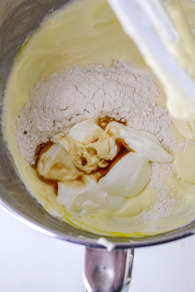 A mixing bowl containing flour, vanilla extract, and sour cream is being combined with a creamy Banana Pudding Cheesecake batter.