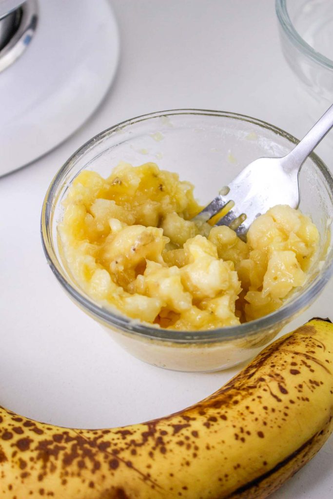 A glass bowl of mashed banana with a fork inside sits next to a ripe, speckled banana on a white surface—perfect for making creamy Banana Pudding Cheesecake.