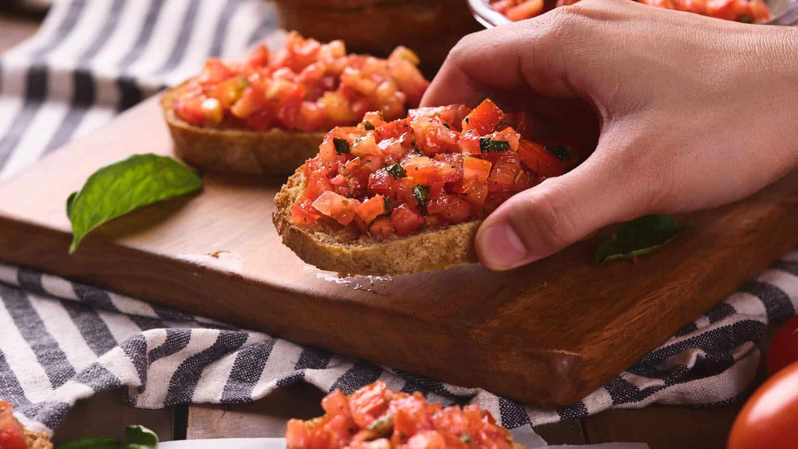 A hand picks up a slice of bruschetta topped with diced tomatoes and herbs from a wooden board—a nod to classic recipes. Other bruschetta pieces and a basil leaf are visible nearby.