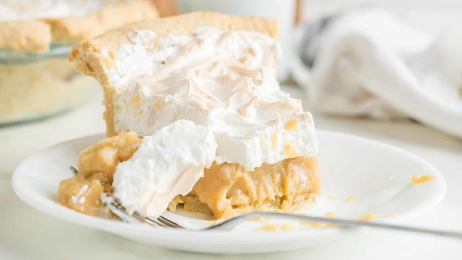 A slice of meringue-topped pie with a creamy filling sits on a white plate; a fork rests in front with some pie on it.