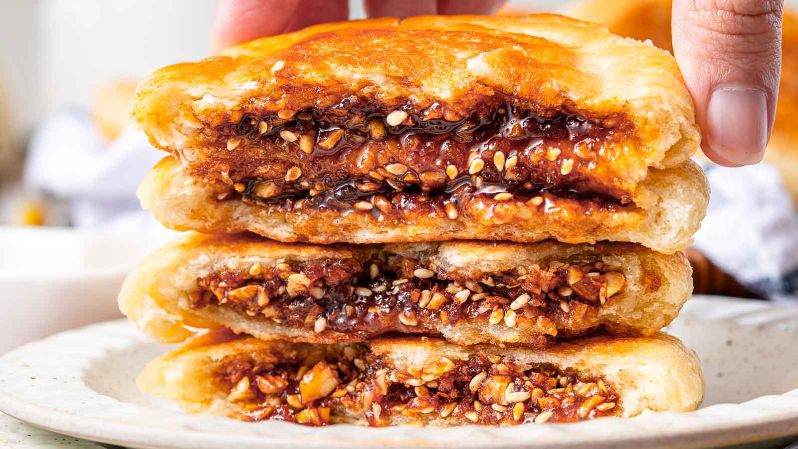 Three halved pastries with a golden, flaky crust are stacked on a plate, revealing a dense filling of chopped nuts and seeds in a sticky, brown syrup. A hand is holding the top piece.