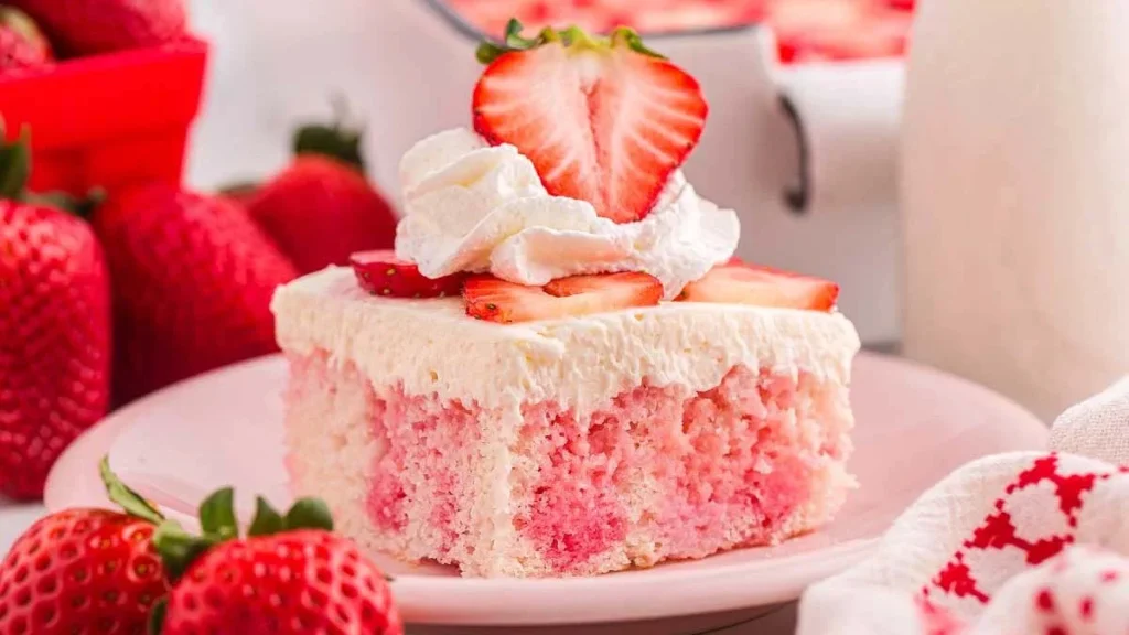 A slice of strawberry poke cake topped with whipped cream and fresh strawberry slices, served on a pink plate with whole strawberries beside it.