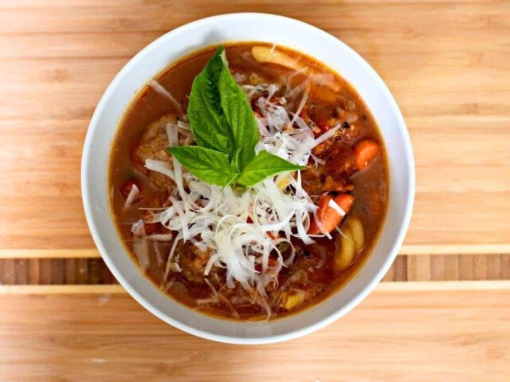 A bowl of tomato-based soup with shredded cheese, sliced carrots, pasta, and a fresh basil garnish, set on a wooden surface—just like one of those timeless recipes passed down through generations.