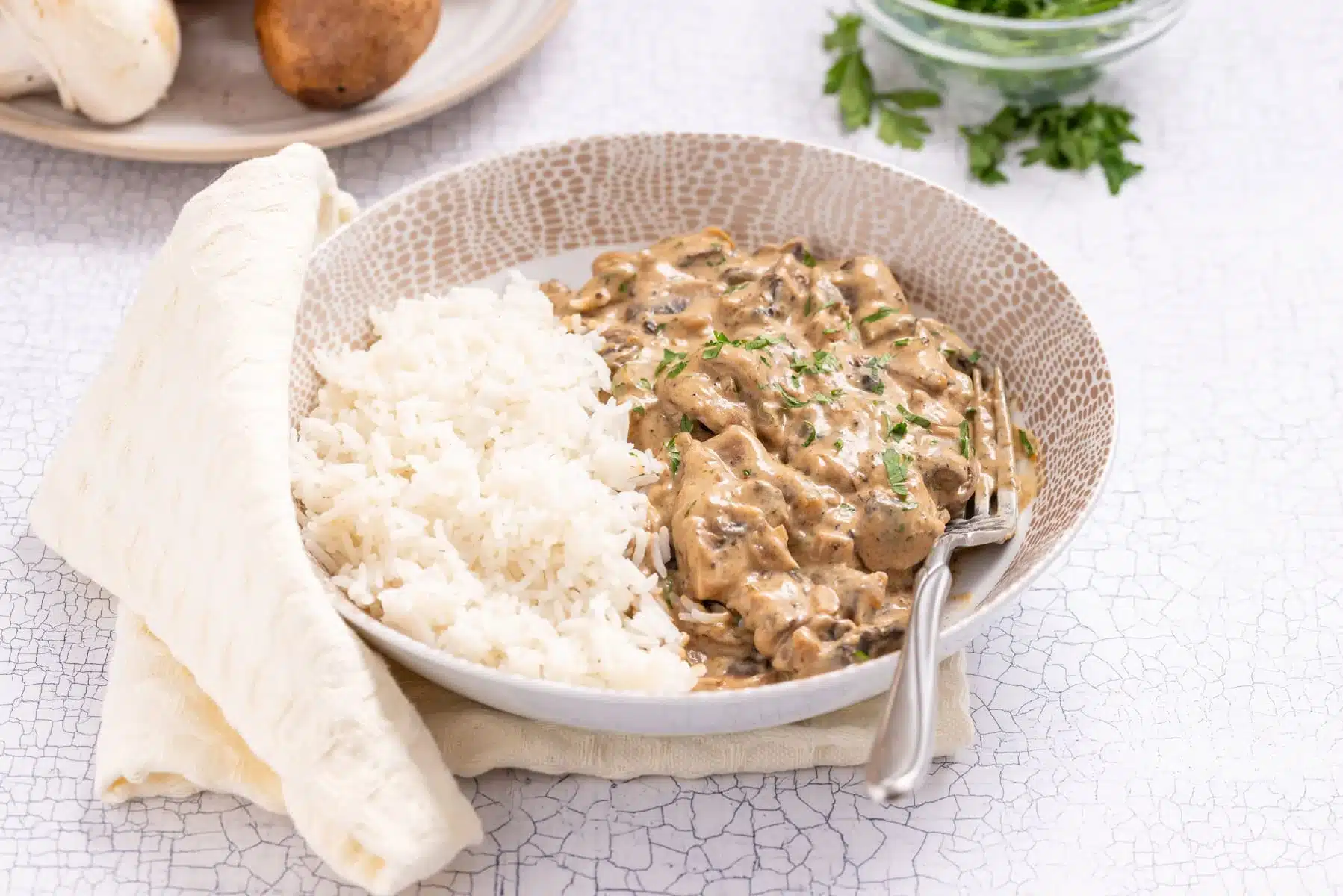 A bowl of white rice and creamy beef stroganoff garnished with herbs, served with a fork and a piece of flatbread on the side.