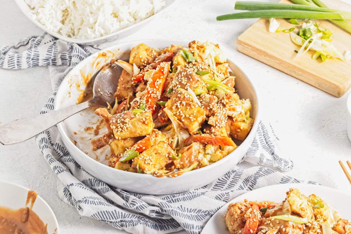 A bowl of tofu and vegetables in a brown sauce, garnished with sesame seeds and green onions, with a bowl of rice and chopped scallions nearby.