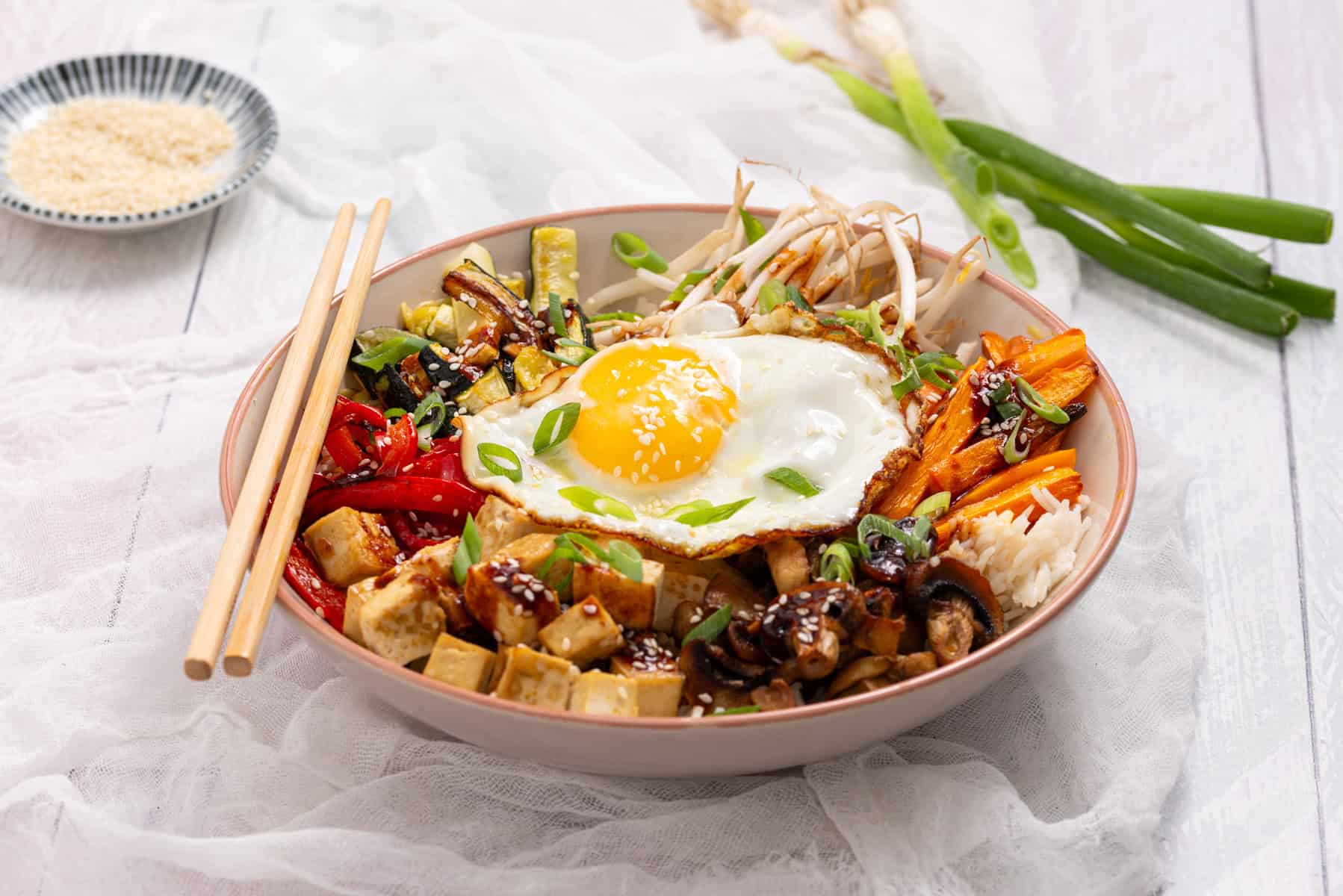A bowl of rice topped with a fried egg, tofu, sliced vegetables, and green onions, with chopsticks resting on the side.