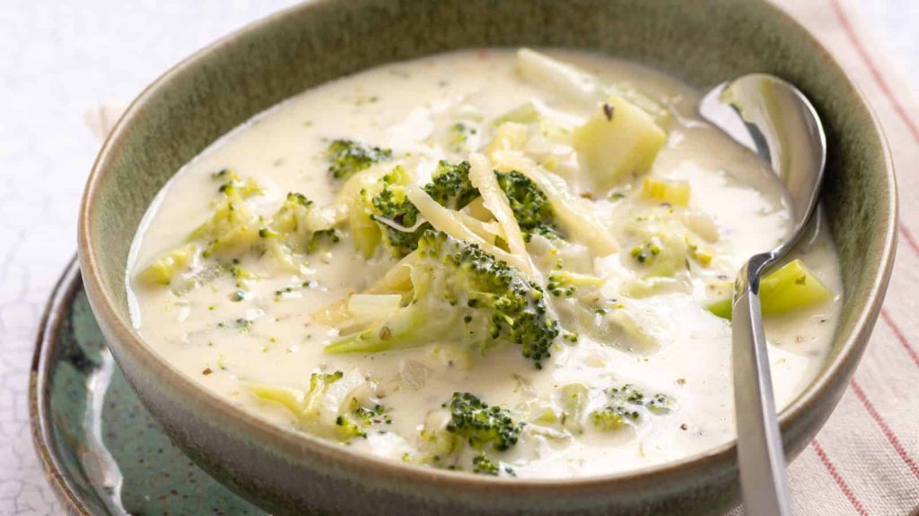 A bowl of creamy broccoli and cheese soup with visible broccoli florets and shredded cheese, served with a spoon on the side—a comforting classic inspired by timeless recipes.