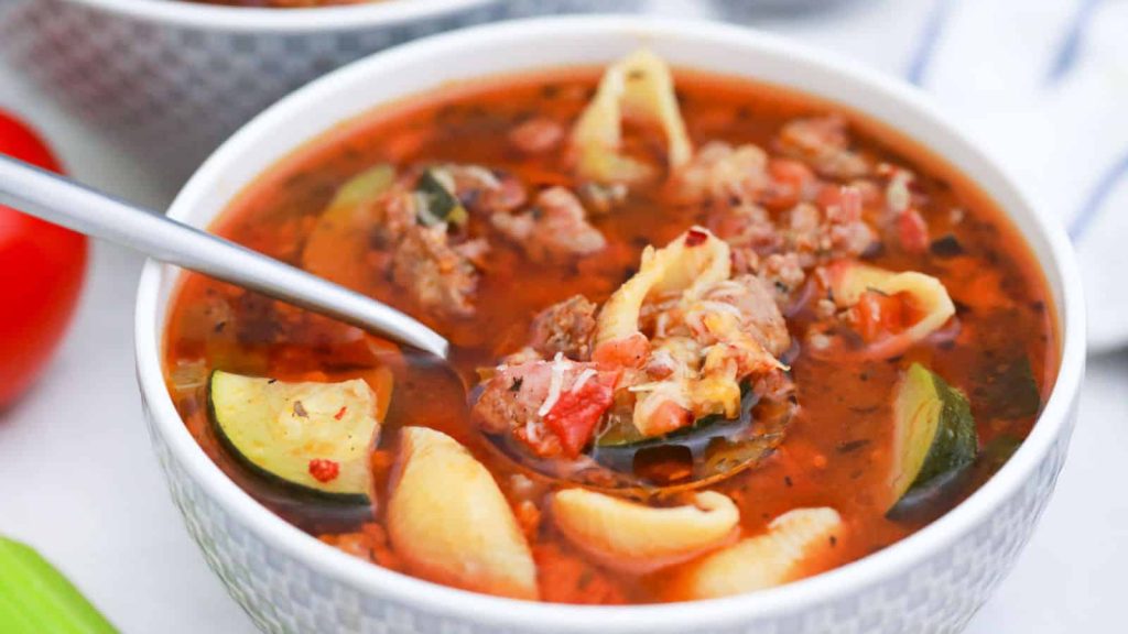 A bowl of tomato-based soup with ground meat, pasta shells, zucchini slices, and herbs—a comforting dish inspired by classic family recipes—with a spoon resting inside the bowl.