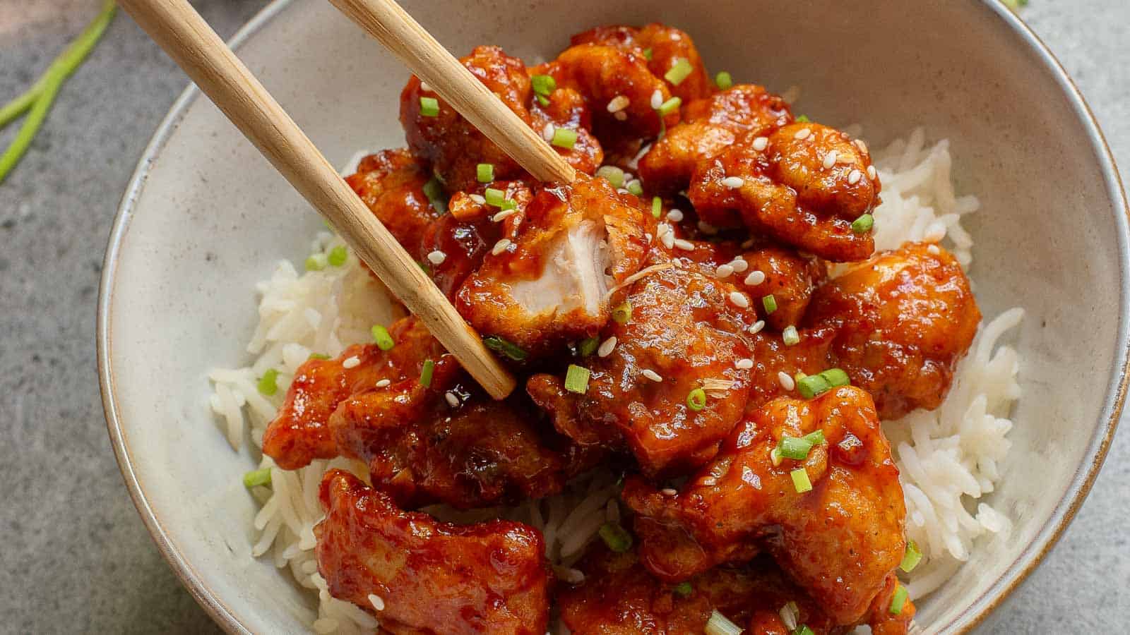 A bowl of white rice topped with glazed fried chicken pieces, garnished with chopped green onions, with chopsticks picking up a piece.