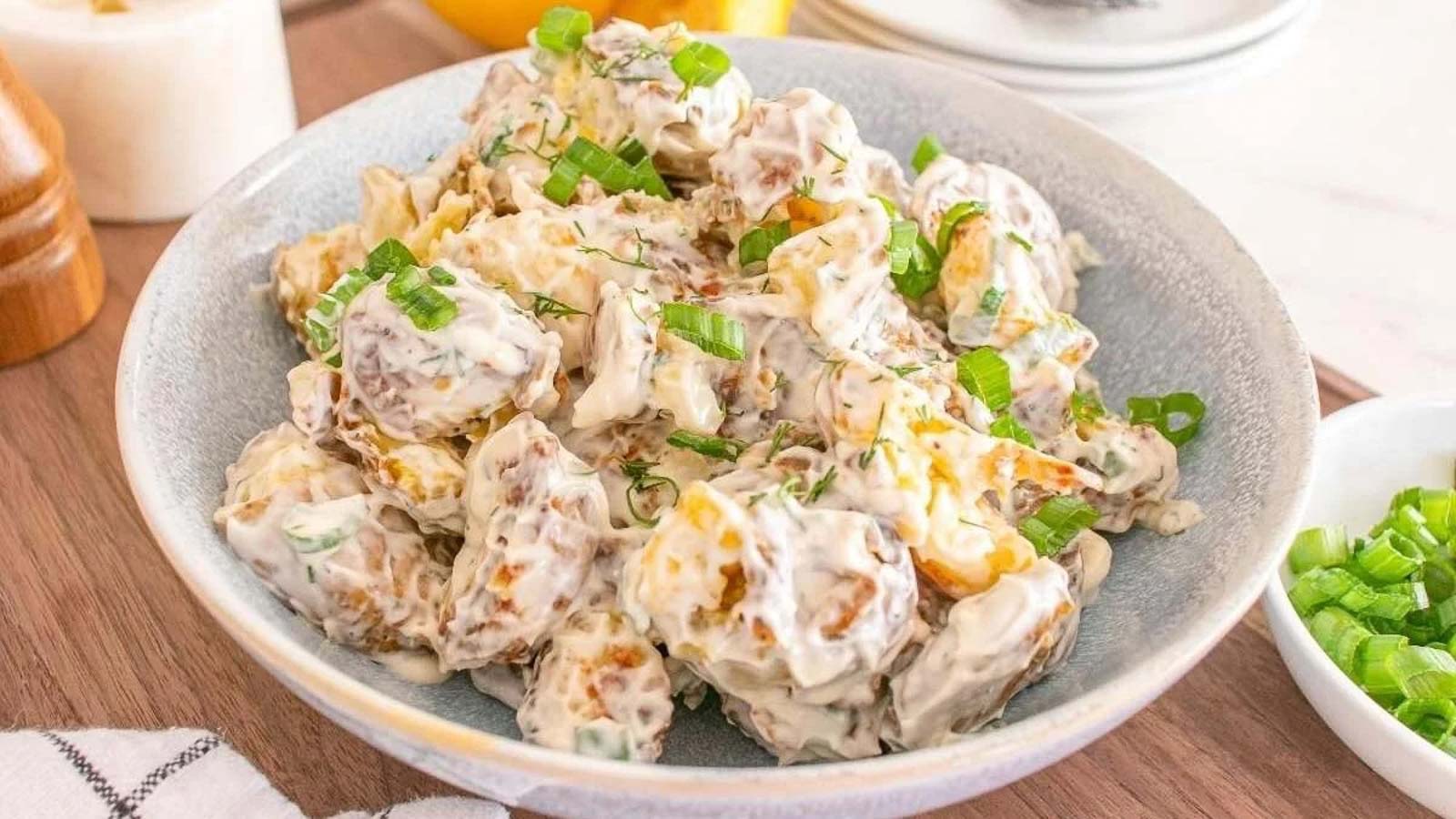 A bowl of roasted potatoes coated in a creamy sauce, garnished with chopped green onions, sitting on a wooden table.