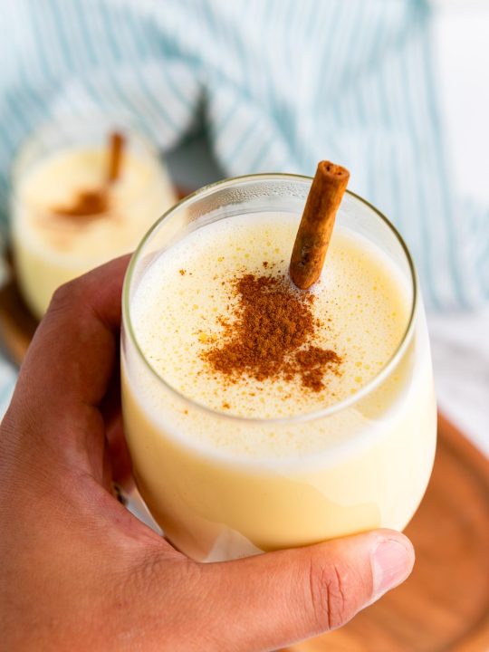 A hand holds a glass of homemade eggnog topped with ground cinnamon and a cinnamon stick, with another glass on a table in the background—perfect for showcasing your favorite eggnog recipe.