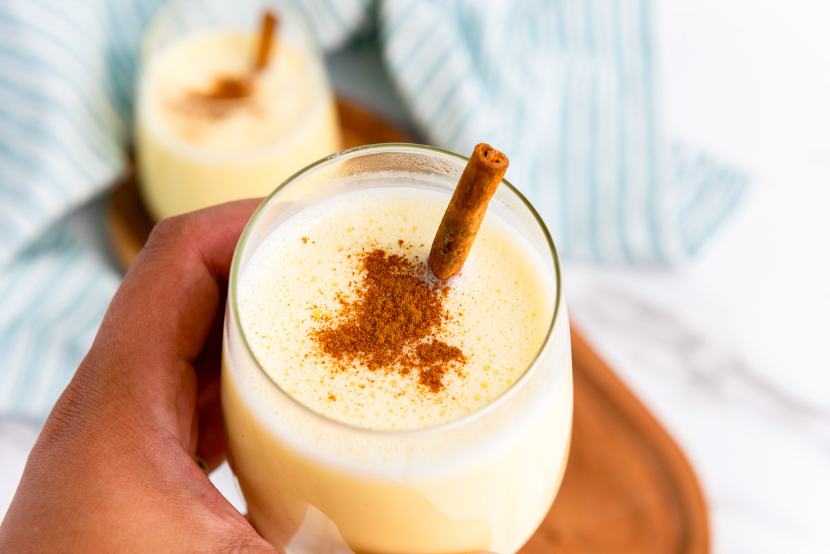 A hand holding a glass of homemade eggnog topped with ground cinnamon and a cinnamon stick, with another glass and a striped cloth in the background—perfect inspiration for your next homemade holiday drinks or eggnog recipe.