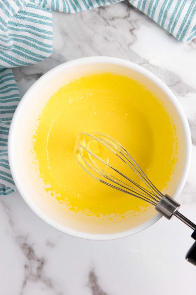 A metal whisk in a white bowl with yellow liquid batter, perfect for whipping up a delicious homemade eggnog recipe, sits on a marble surface next to a blue and white striped cloth.