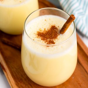 A glass of creamy yellow homemade eggnog topped with ground cinnamon and garnished with a cinnamon stick, placed on a wooden tray—a classic homemade holiday drink.