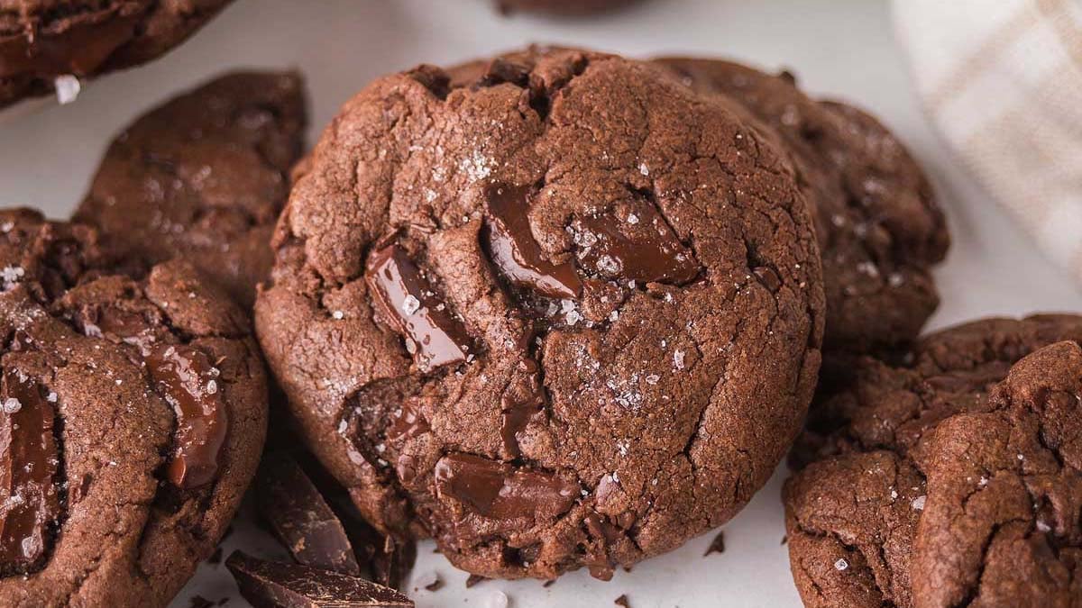 Close-up of several chocolate cookies with chunks of dark chocolate and a sprinkling of sea salt on a white surface.