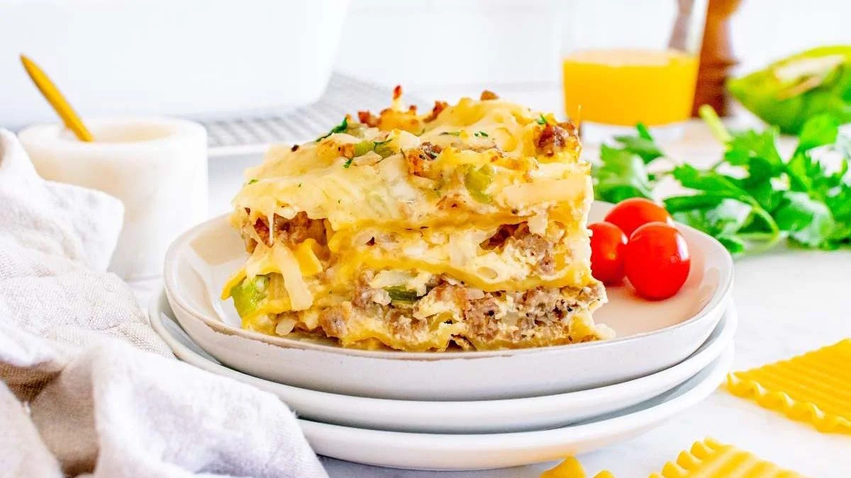 A slice of lasagna with layers of pasta, cheese, and meat sits on a white plate, garnished with herbs, with tomatoes and greens in the background.