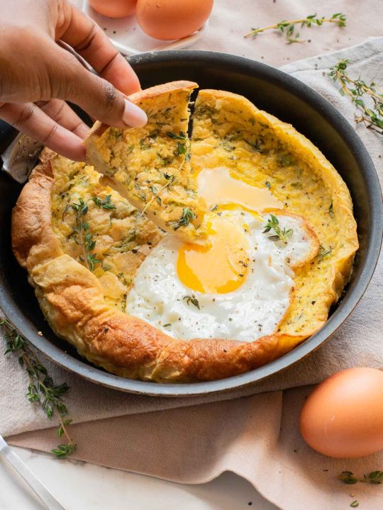 A hand lifts a piece of savory Dutch baby pancake, brimming with herbs and topped with a fried egg, from a skillet set beside fresh eggs and sprigs of thyme.