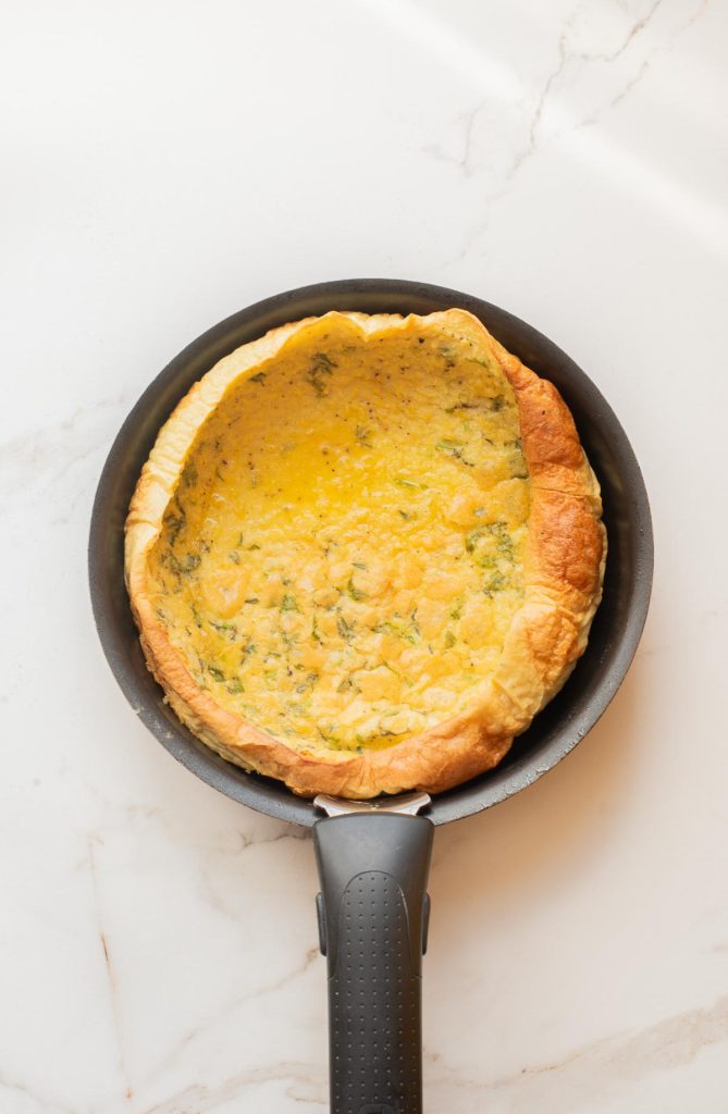A fluffy, savory Dutch baby omelette with herbs, baked to perfection in a nonstick frying pan on a white marble surface.