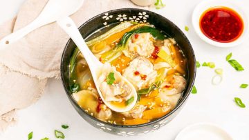 A bowl of wonton soup with vegetables, garnished with green onions and sesame seeds, served with a white ceramic spoon. A small dish of red chili oil is on the side.